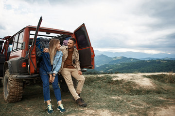 Joyful young couple of travelers resting near their car while enjoying their weekend in the mountains