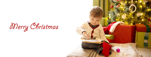 Cute little boy reading a book in front of the christmas tree at home with copy space for text