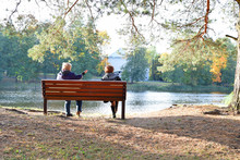 Two Mature Women Sitting On Wooden Bench And Talking Each Other Next To Lake In Park In Autumn Day.               