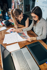 Wall Mural - young female graphic designers sitting at desk with laptop, color charts and some sketches discussin