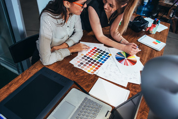 Two female architects working together using color swatches sitting at desk with laptop, graphic tablet in design studio