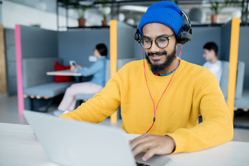 positive satisfied middle-eastern app developer in headphones sitting at table and using laptop whil