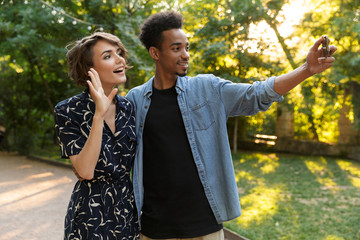 Poster - Smiling young multiethnic loving couple standing posing in park outdoors hugging take a selfie by phone.