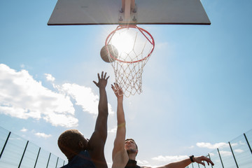 Low angle view of two young men playing basketball and jumping by hoop against blue sky lit by sunlight, copy space