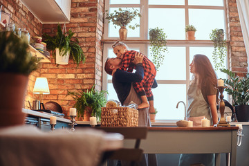 Wall Mural - Happy dad holds his little girl on hands while mom standing near in loft style kitchen at morning.