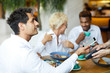 © pressmaster - Happy handsome young man in whit shirt sitting at table in restaurant and looking at waitress while using smartphone for contactless payment