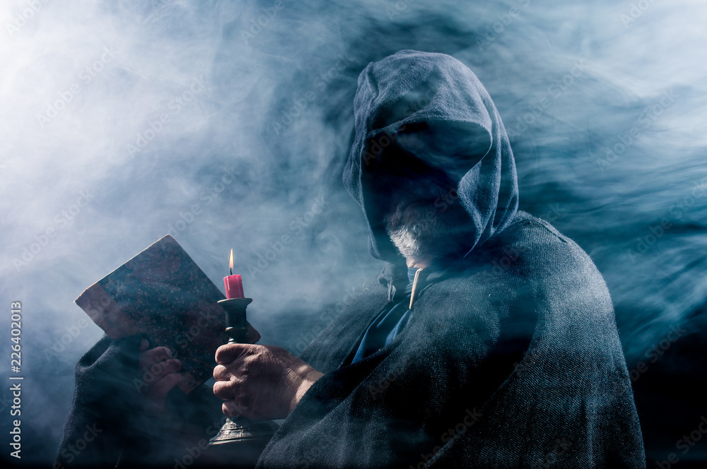 a mysterious figure holds a book and a candle in his hands - obrazy, fototapety, plakaty 