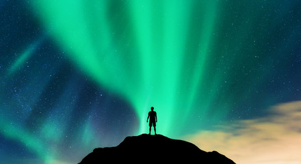 Wall Mural - Aurora and silhouette of alone standing man on the mountain peak. Lofoten islands, Norway. Aurora borealis and young man. Sky with stars and green polar lights. Night landscape with northern lights