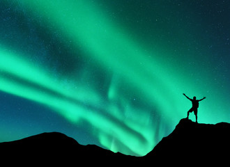 Wall Mural - northern lights and silhouette of standing man with raised up arms on the mountain peak in norway. a