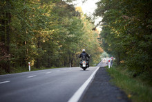 Back View Of Motorcyclist In Black Leather Jacket And White Helmet Riding Cruiser Motorbike Along Hilly Road Between Tall Green Trees. Active Lifestyle, Love To Adventures Concept.