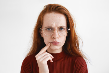 Picture of puzzled young red haired woman in spectacles frowning, having suspicious look, touching lips. Beauitful pensive ginger girl being frustrated with something. Human facial expressions