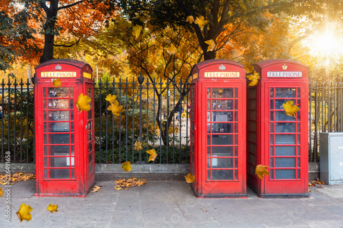 Fotografie, Obraz Klassisch, rote Telefonzellen aufgereiht vor einem Park im Herbst in London