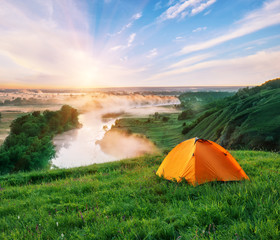 Wall Mural - orange tourist tent on a hill with green grass above river