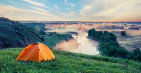Wall Mural - orange tourist tent on hill with green grass above the small riv