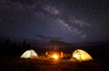 Camping In Mountains At Night. Bright Bonfire Burning Between Two Hikers, Boy And Girl Sitting Opposite Each Other Near Illuminated Tents Under Beautiful Evening Starry Sky And Milky Way