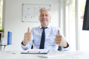 Shot of senior businessman giving two thumbs up while sitting at office desk and looking at camera.