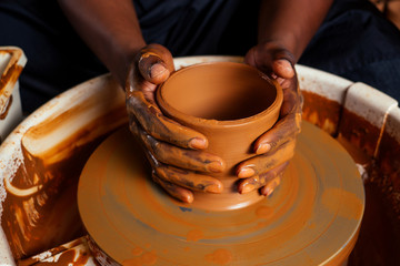 mixed race afro male potter with black apron and stylish dark shirt sitting at workshop table potter's wheel , applying glaze on handmade clay brown pot vase