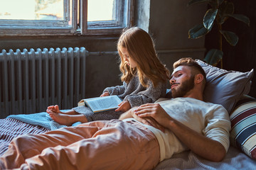 Wall Mural - Handsome young father and daughter reading storybook together while lying on bed.