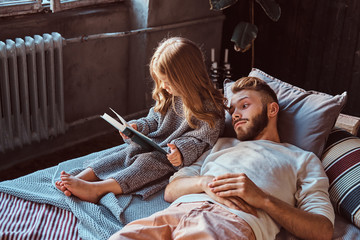 Wall Mural - Handsome young father and daughter reading storybook together while lying on bed.