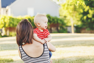 Wall Mural - Group portrait of Caucasian mother with baby boy in red t-shirt in field meadow outside. Female parent holding little happy smiling son child in summer park on sunset. Happy parenting lifestyle.