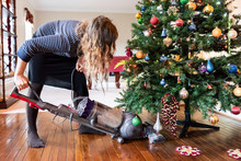 Young Woman Cleaning With Vacuum Cleaner, Vacuuming Under Christmas Tree Needles With New Years Ornaments On Hardwood Wooden Floor