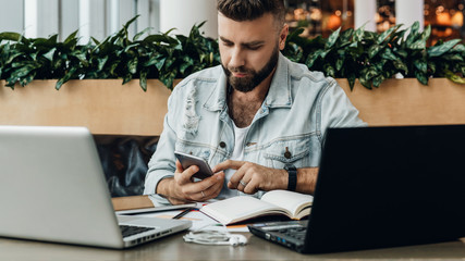 Hipster man sits in cafe, uses smartphone, works on two laptops. Businessman reads an information message in phone.