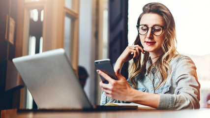 Wall Mural - Young businesswoman is sitting in coffee shop at table in front of computer and notebook,using smartphone. Social media.