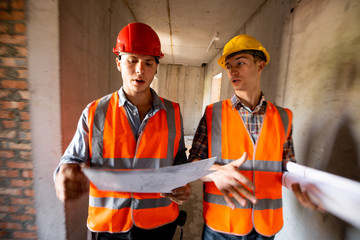 Two men dressed in orange work vests and helmets work with construction documentation inside the building under construction