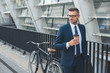 © LIGHTFIELD STUDIOS - handsome businessman in suit and eyeglasses holding paper cup and looking away while standing with bike on street