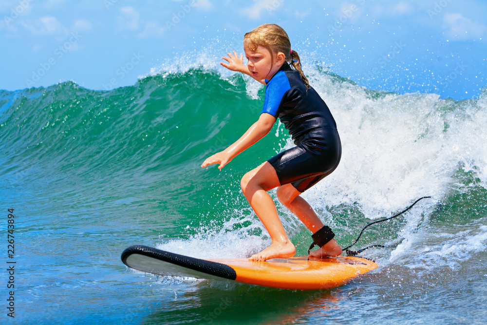 Happy baby girl - young surfer ride on surfboard with fun on sea waves. Active family lifestyle, kids outdoor water sport lessons and swimming activity in surf camp. Summer vacation with child. - obrazy, fototapety, plakaty 