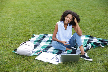 Poster - Smiling young african girl sitting on grass outdoors in park chatting by phone.