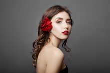 Woman With A Big Red Flower In Her Hair. Brown-haired Girl With A Red Flower Posing On A Gray Background. Big Beautiful Eyes And Natural Makeup. Long Curly Hair, Perfect Face
