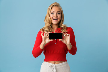 Wall Mural - Portrait of optimistic blond woman 20s wearing red shirt smiling and showing empty screen of cell phone, isolated over blue background in studio