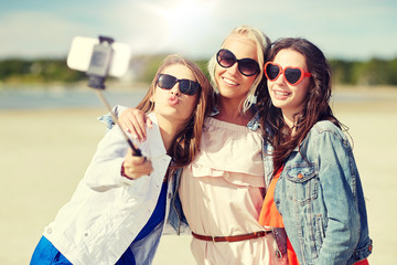 Poster - summer vacation, holidays, travel, technology and people concept- group of smiling young women taking picture with smartphone on selfie stick on beach