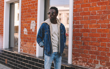 Wall Mural - Fashion african man wearing jeans jacket poses on city street, brick wall background