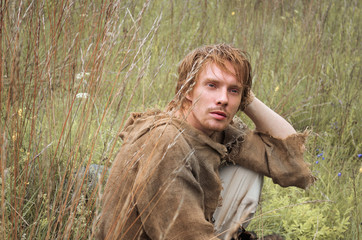 Wall Mural - young shaggy and unshaven guy dressed as a medieval peasant from an old bag thought about something sitting in a field among the yellowed wild herbs