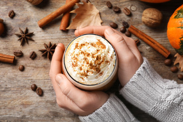 Woman holding glass of tasty pumpkin spice latte on wooden table, flat lay composition