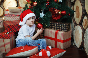 Boy in Christmas hats is sitting with gift boxes in his hands with the joy of a surprise.