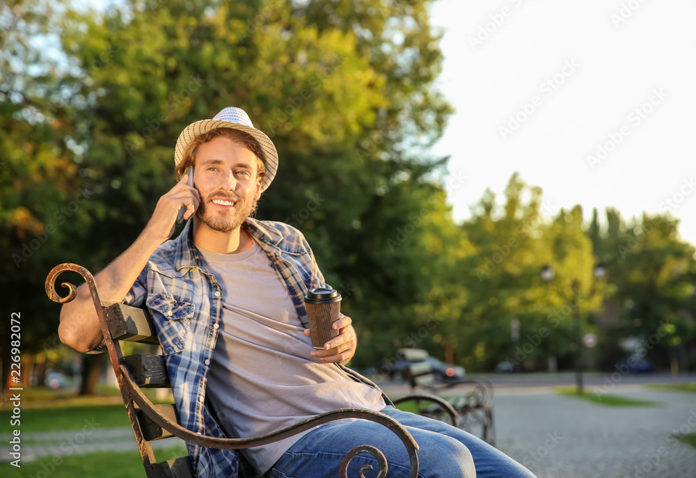 Handsome young man with coffee talking by mobile phone in park