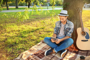 Sticker - Handsome young man with tablet PC resting on plaid in park