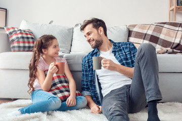 Father and little daughter at home sitting on floor drinking hot chocolate joyful