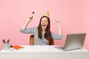 Wall Mural - Cheerful girl with closed eyes in birthday party hat with playing pipe screaming celebrating while sit work at desk with pc laptop isolated on pink background. Achievement business career. Copy space.