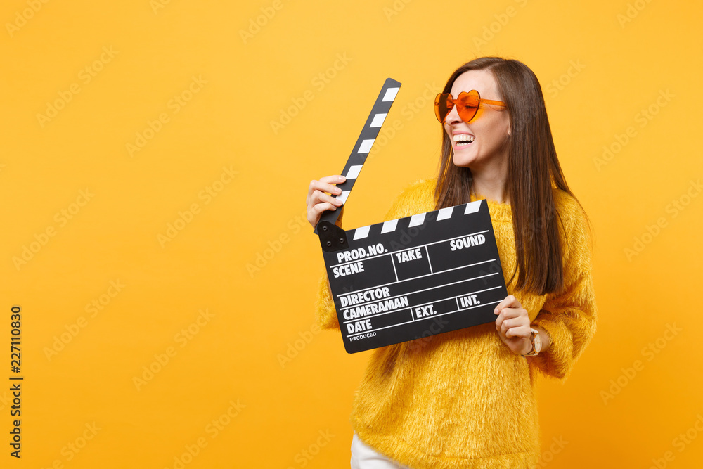 Laughing young woman in orange heart eyeglasses looking aside and holding classic black film making clapperboard isolated on yellow background. People sincere emotions, lifestyle. Advertising area. - obrazy, fototapety, plakaty 
