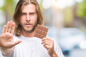 Poster - Young handsome man with long hair eating chocolate bar over isolated background with open hand doing stop sign with serious and confident expression, defense gesture