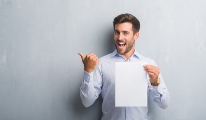 Sticker - Handsome young man over grey grunge wall holding blank paper sheet contract pointing and showing with thumb up to the side with happy face smiling