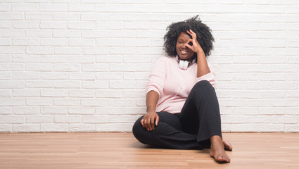 Canvas Print - Young african american woman sitting on the floor wearing headphones doing ok gesture with hand smiling, eye looking through fingers with happy face.