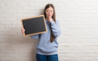 © Krakenimages.com - Young Chinese woman over brick wall holding blackboard serious face thinking about question, very confused idea