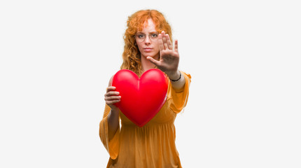 Sticker - Young redhead woman in love holding red heart with open hand doing stop sign with serious and confident expression, defense gesture