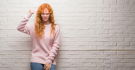 Canvas Print - Young redhead woman standing over brick wall angry and mad raising fist frustrated and furious while shouting with anger. Rage and aggressive concept.