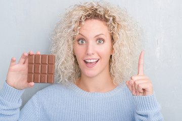 Young blonde woman over grunge grey wall eating chocolate bar surprised with an idea or question pointing finger with happy face, number one